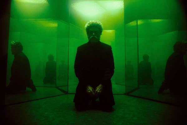 A man kneeling in a mirrored chamber illuminated by vibrant green light, creating multiple reflections.