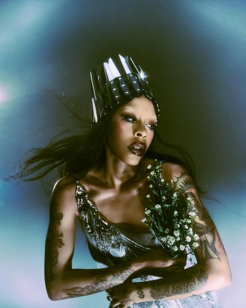 Woman with crown poses indoors with flower bouquet. Person displays tattoos and wears metallic top with dramatic eye makeup in portrait.