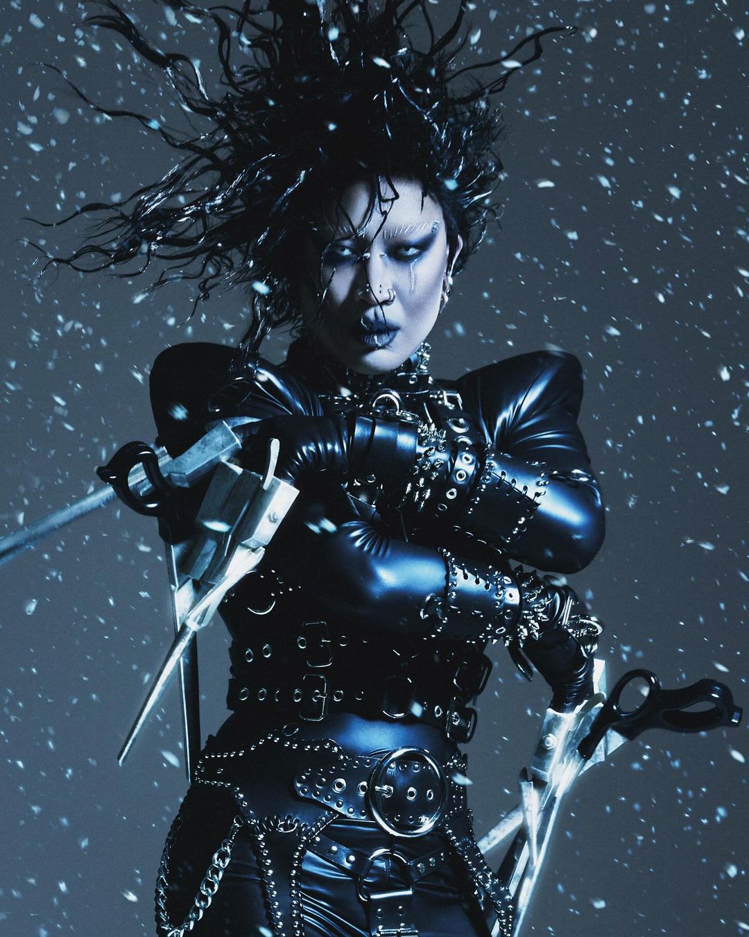 Person in elaborate black leather costume, scissors-hands, pale face, dark makeup, teased black hair, and silver studio-style lighting with falling snow.