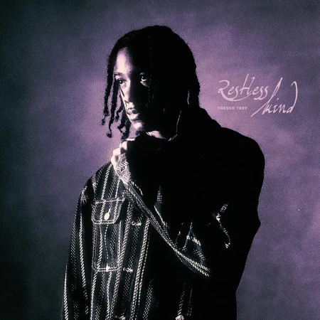Portrait of a young adult male with dreadlocks wearing a denim jacket and rings, against a purple studio background.