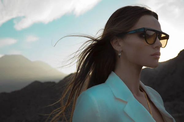 Adult woman with sunglasses poses outdoors, with mountain range and bright sky in background. Fashion, beauty, portrait.