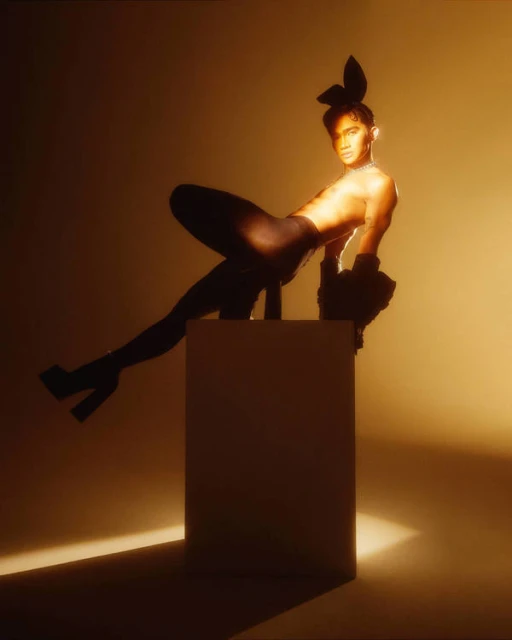 Person with rabbit ears posing in a studio with warm lighting. The person is wearing gloves, black tights and platform heels.