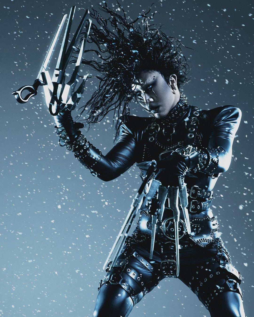 Person dressed as Edward Scissorhands in black studded suit, holding scissor-like hands against a grey background with snow falling.