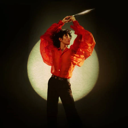 Person with dark curly hair wearing a red sheer blouse holding a silver dagger above head in a spotlight