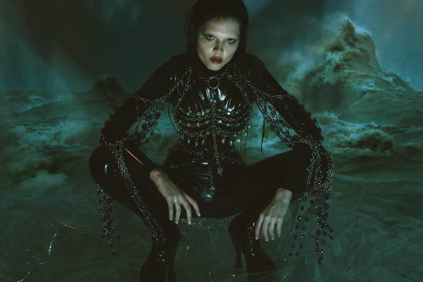 A woman in a black latex outfit and chains squats against a backdrop of a stormy sea with crashing waves in a moody, dark setting.