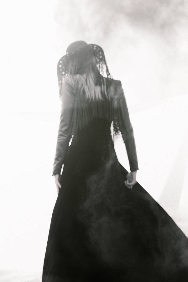 Woman in a long black dress and ornate head covering stands in bright white light, viewed from behind, highlighting fashion and style.