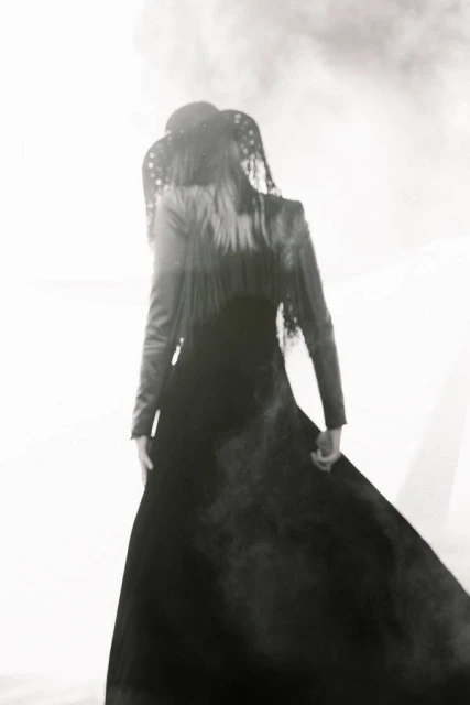 Woman in a long black dress and ornate head covering stands in bright white light, viewed from behind, highlighting fashion and style.