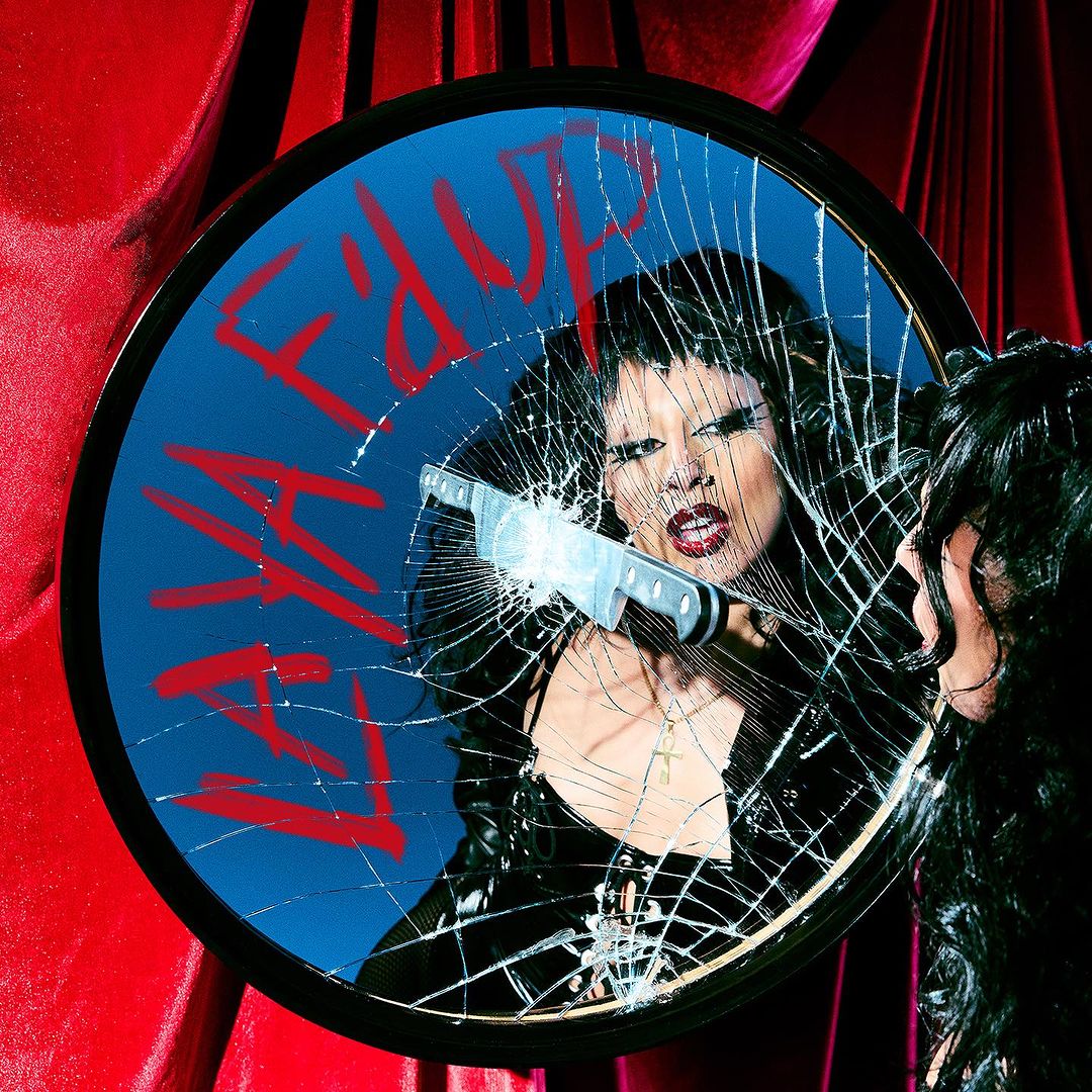 Reflection in shattered mirror of adult female with dark hair and red lips, wearing a black top, surrounded by red fabric.