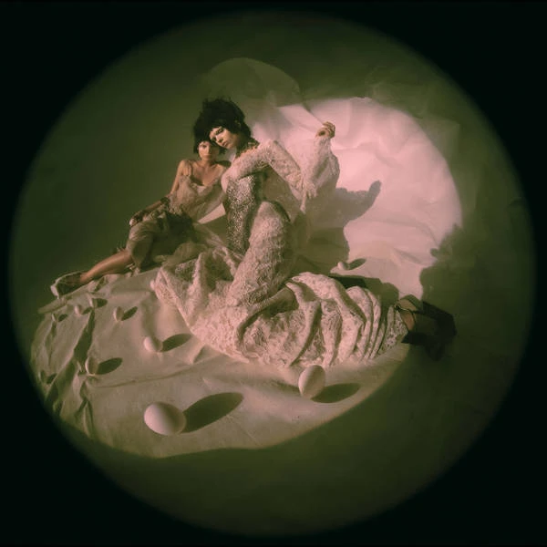 Two people pose on a white sheet with eggs under spotlight in a studio setting, wearing couture fashion against a dark backdrop.