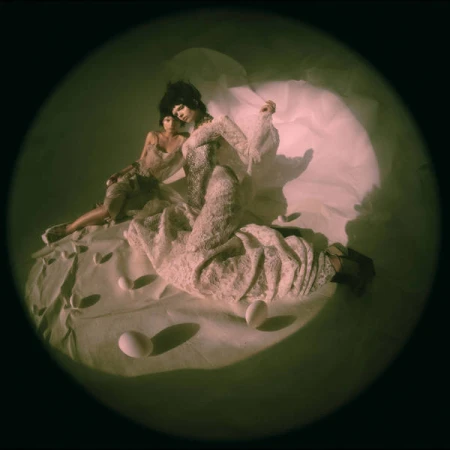 Two people pose on a white sheet with eggs under spotlight in a studio setting, wearing couture fashion against a dark backdrop.