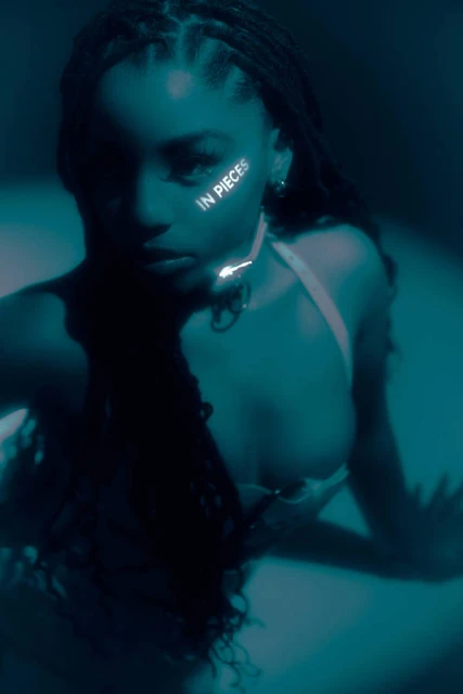 Close-up studio portrait of a woman with braided hair wearing a halter top, with the words 'IN PIECES' visible on her face.
