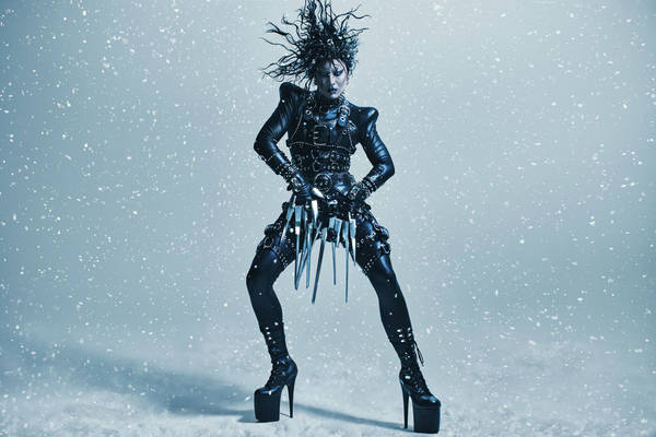 Person in elaborate black costume with blade-like details poses against a cool-toned backdrop with bokeh-like light effect.