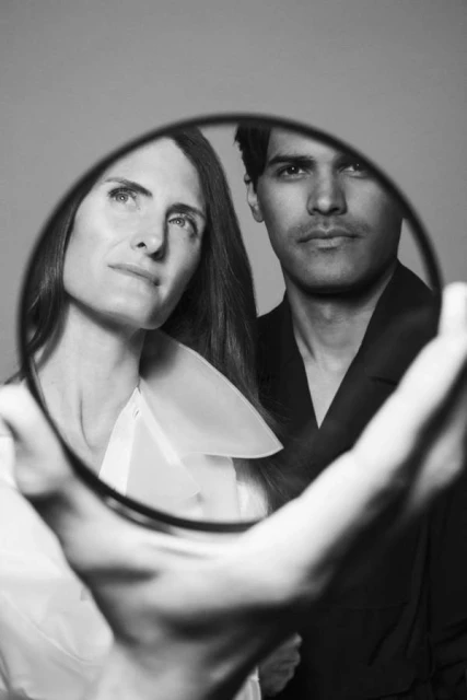 Black and white portrait of a woman in a white collared shirt and a man in a black suit, viewed through an oval mirror held by a person.