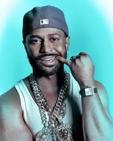 Portrait of a man wearing a cap, white tank top, layered silver chains, and grillz on teeth against a blue background.