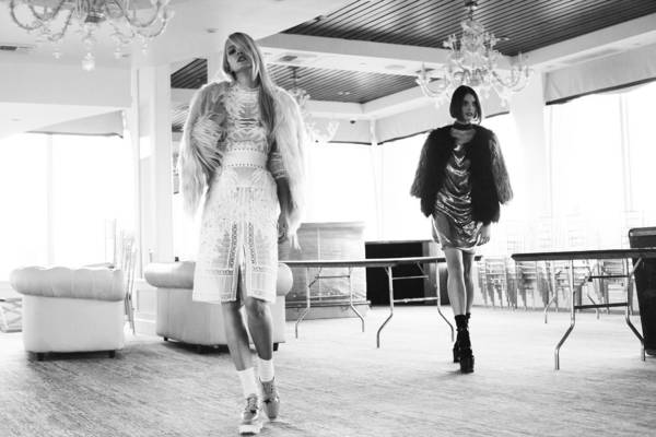 Two female models in fashionable dresses walk in a brightly lit indoor space with minimalist decor and modern chandeliers, black and white.