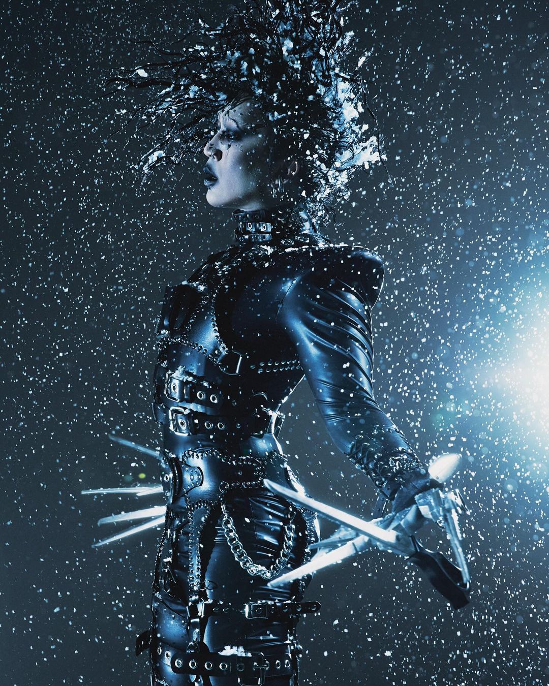 Side profile of a person in a stylized costume with silver scissor hands, hair, and studded black clothing, with snow falling around them.