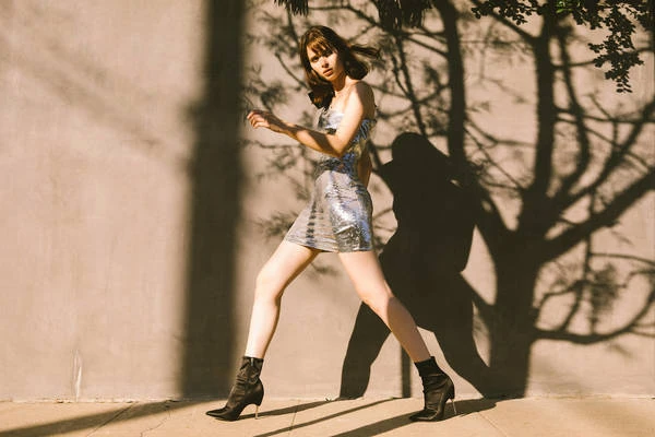 Young woman in a silver dress and heeled boots walks along a sidewalk in the sunlight with tree shadows cast on a wall behind her.