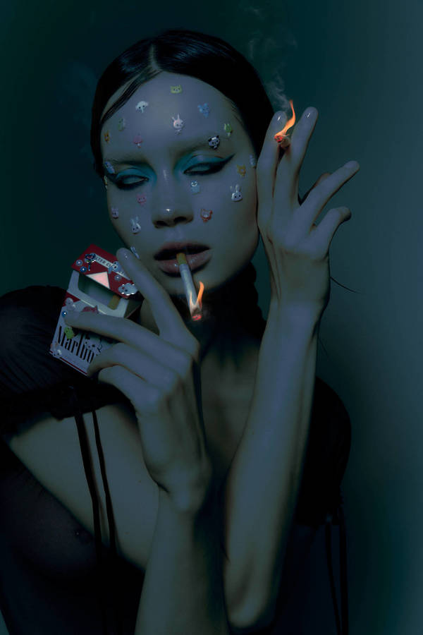 Portrait of a woman with face stickers, blue eyeshadow, holding a cigarette and a pack, photographed by Brian Ziff