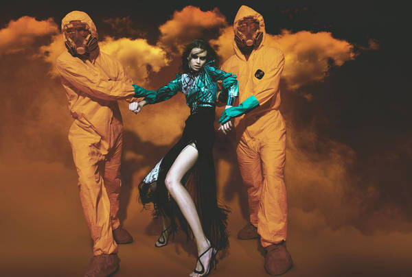 Fashion shot of a model in a dress between two people in hazmat suits against a hazy background with orange and black tones.