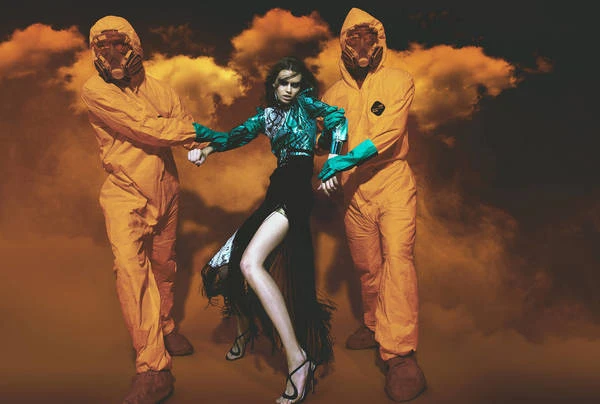 Fashion shot of a model in a dress between two people in hazmat suits against a hazy background with orange and black tones.