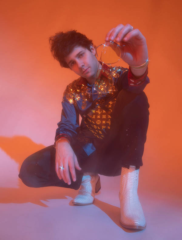 Portrait of a young man squatting in a photo studio with a shiny shirt, black pants, and cowboy boots against an orange backdrop.