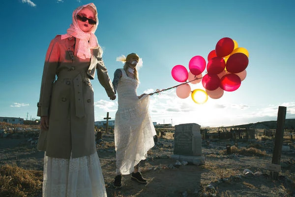 Two women in vintage fashion hold red balloons in a sunny cemetery against a blue sky, creating a striking and somewhat surreal image.