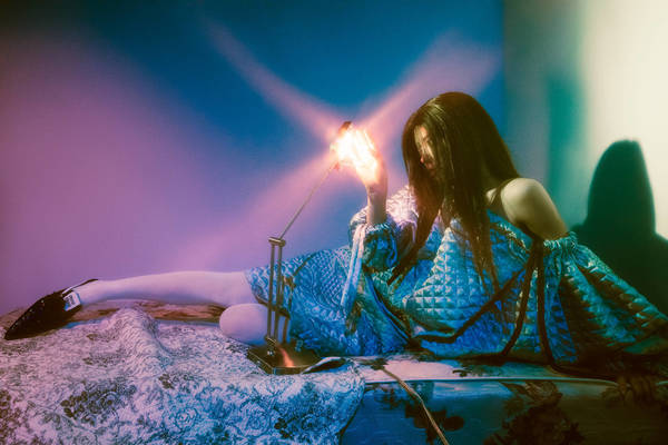 Woman in blue dress sits on patterned bed with lamp creating light play on wall. Shadow and light create dramatic effect.