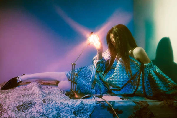 Woman in blue dress sits on patterned bed with lamp creating light play on wall. Shadow and light create dramatic effect.