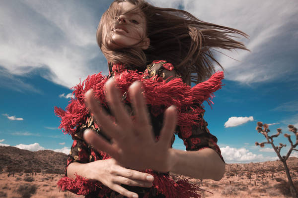 Woman in a patterned dress with red fringed sleeves poses in a desert landscape under a bright blue sky with scattered clouds.