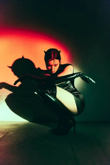 Woman in a black dress and thigh-high boots crouches in a photo studio with red and green lighting, casting a shadow on the wall.