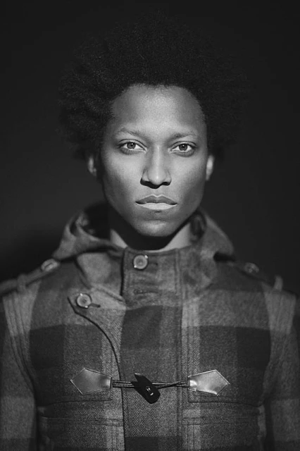 Headshot of an African-American adult male with a neutral expression wearing a patterned hooded coat in a dark indoor environment with soft lighting.