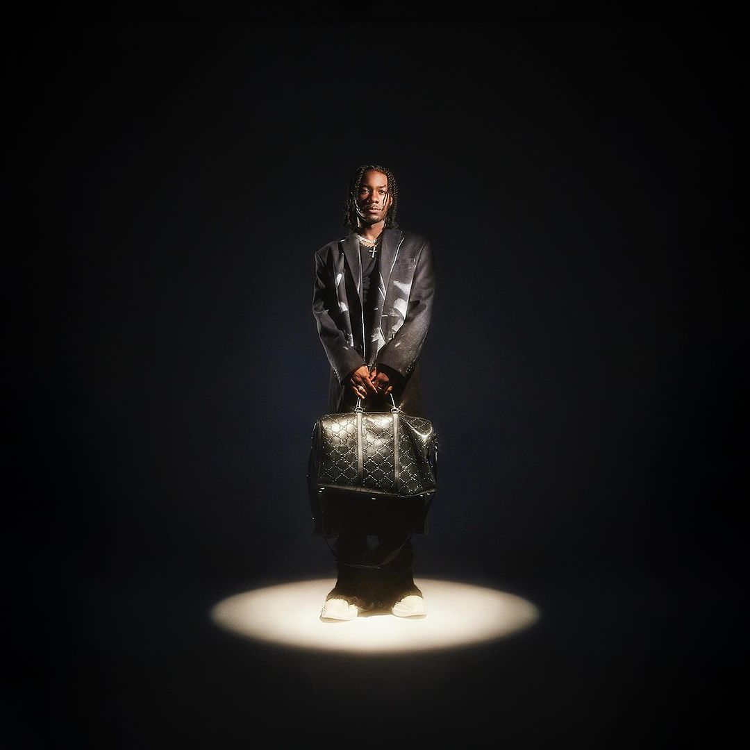 Stylish man stands in spotlight wearing a blazer, pants, and sneakers, holding a patterned travel bag in a dark studio setting.