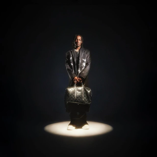 Stylish man stands in spotlight wearing a blazer, pants, and sneakers, holding a patterned travel bag in a dark studio setting.