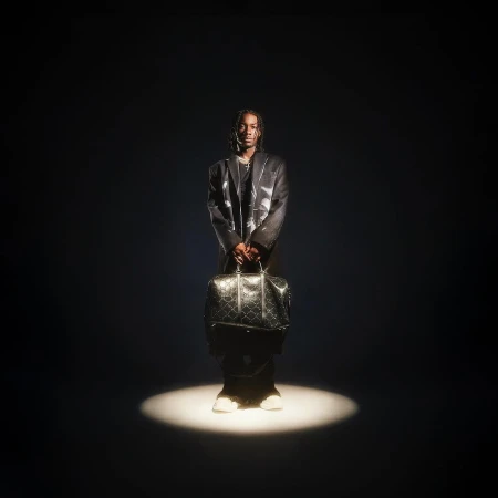 Stylish man stands in spotlight wearing a blazer, pants, and sneakers, holding a patterned travel bag in a dark studio setting.