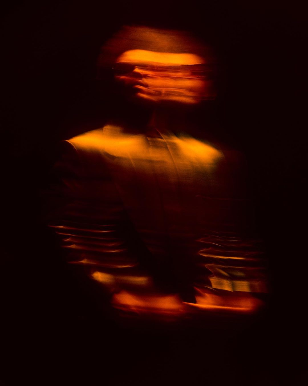 Abstract, motion-blurred silhouette of a person with bright orange light on their face, set against a dark, indistinct background.