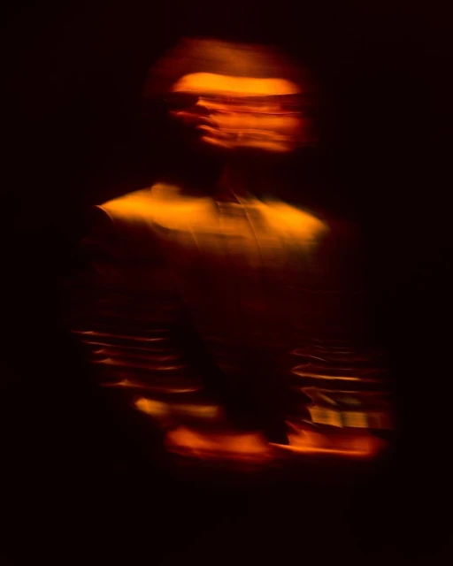 Abstract, motion-blurred silhouette of a person with bright orange light on their face, set against a dark, indistinct background.