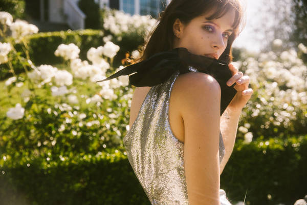 Portrait of a fair-skinned woman with bangs in a silver dress and black bow, posing in front of white flowers and foliage outdoors.
