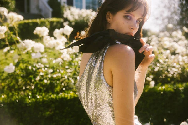 Portrait of a fair-skinned woman with bangs in a silver dress and black bow, posing in front of white flowers and foliage outdoors.