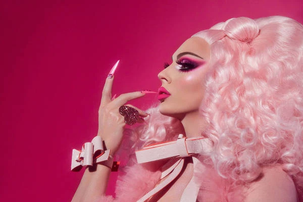Profile view of a drag queen with heavy pink makeup, long pink nails, pink ring, pink wig, and pink furry wrap on a pink backdrop.