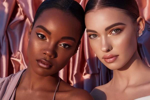 Close-up of two women with professional makeup, clear skin, and neutral expressions, posing in front of a textured pink fabric background.