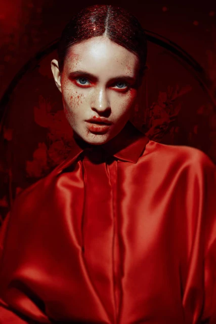 Close-up portrait of a fair-skinned adult woman with red glitter makeup and a red satin shirt against a dark red textured background.