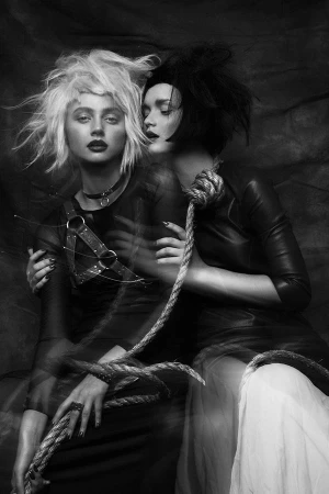 Black-and-white portrait of two women with dramatic hair and makeup, sharing a length of rope, against a textured backdrop.