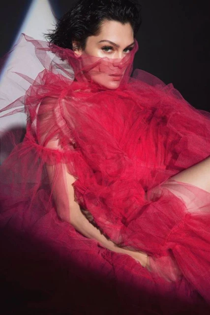 Adult female in red tulle gown and short dark hair poses in a studio with a soft spotlight in a dark background. Fashion editorial shot.