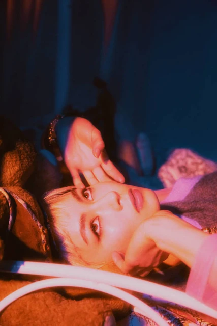 Close-up portrait of a fair-skinned adult female laying down with hands near her face under colorful lighting in an indoor setting.