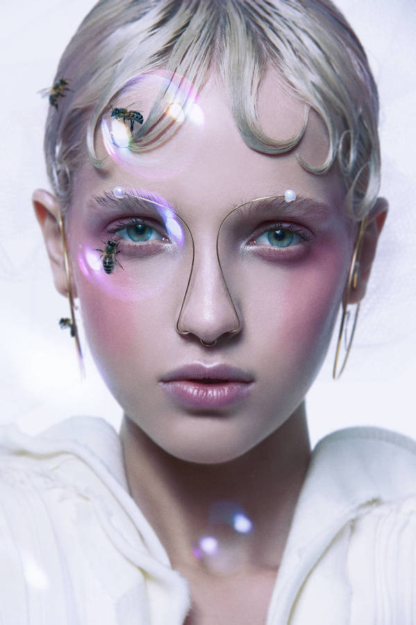 Close-up portrait of a model with pale skin, light blonde hair, and pink makeup, adorned with bees and geometric wire framing the face.