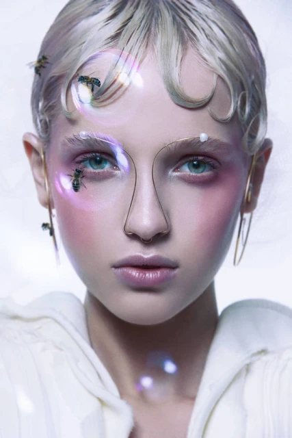 Close-up portrait of a model with pale skin, light blonde hair, and pink makeup, adorned with bees and geometric wire framing the face.