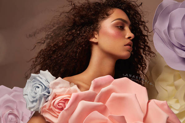 Close-up of a woman with curly hair and pink blush, adorned with pastel-colored roses, against a muted brown backdrop in a beauty campaign.