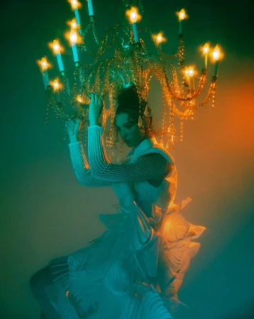 Woman poses under ornate chandelier in artistic studio shot with diffused, colorful lighting; she wears textured top and flowing white skirt.