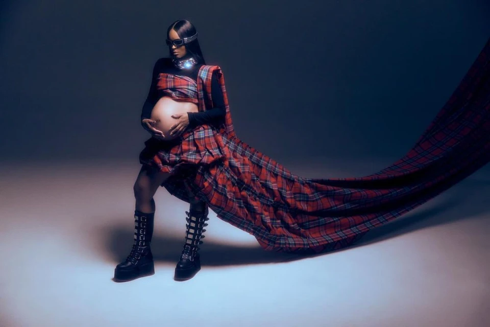 Pregnant woman in red plaid dress and black platform boots poses in a studio with hands on stomach and a flowing cape.