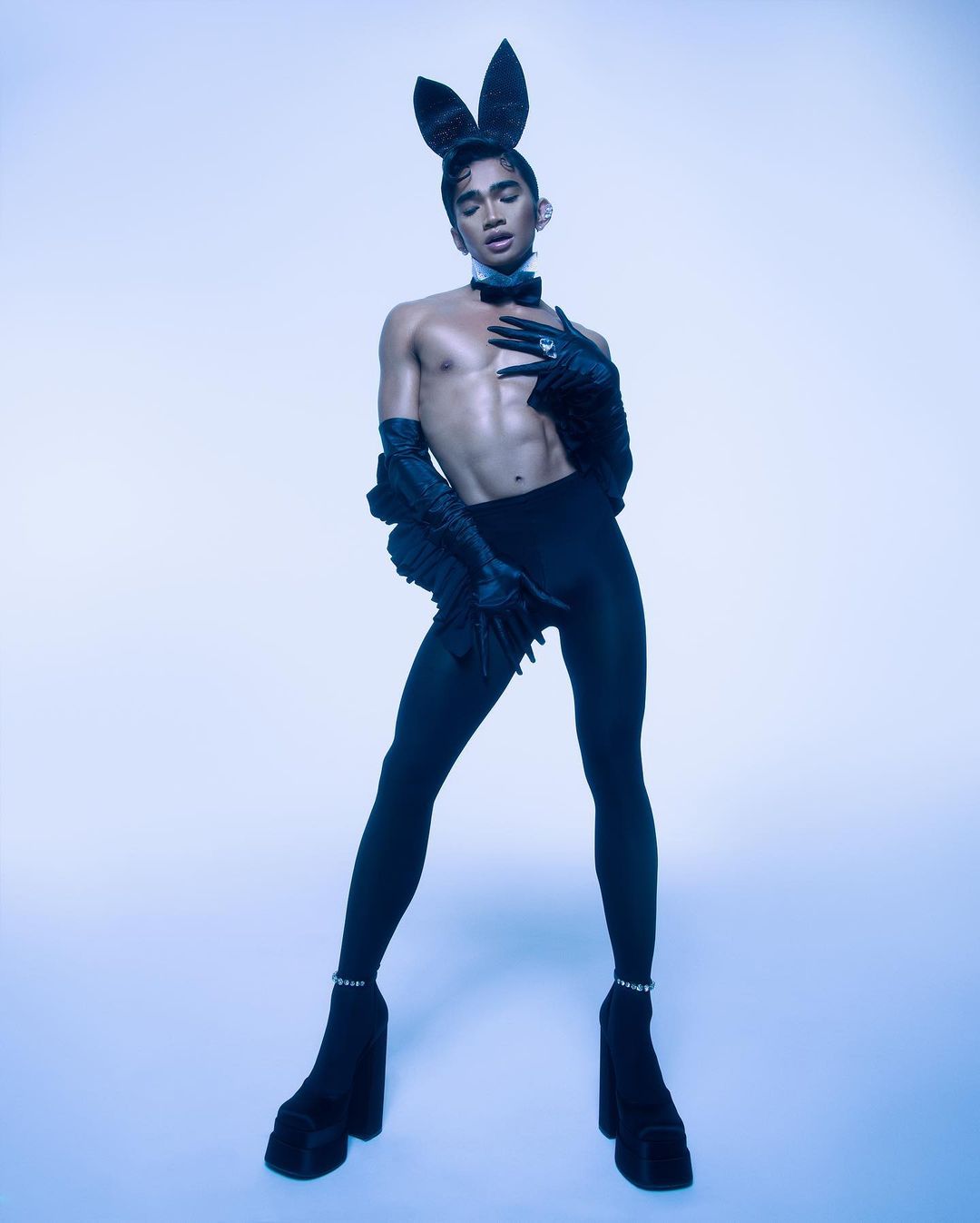 An adult male poses in a studio wearing a bunny costume with high platform boots, black tights, and a bow tie against a light blue background.